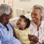 Grandparents Sitting On Sofa At Home Playing With Baby Granddaughter