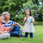 Asian cute granddaughter making bubbles and attractive happy grandparents look at her in the public garden at vacation day or holiday. Family Concept.