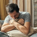 Depressed man sitting on living room sofa and hugging pillow