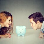 Finances in divorce concept. Wife and husband can not make settlement holding piggy bank sitting at table looking at each other with hatred