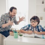 Photo of aggressive dad screaming at his small son who does not understand the material and home task he is very terrified sitting at the table trying to concentrate read textbook