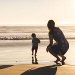 Full length of woman crouching behind toddler son running on shore at beach while enjoying sunset