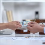 Two hands exchanging cash over a desk with papers and rings visible.