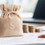 Money bag on desk with coin stacks and documents, symbolizing finance or economy.